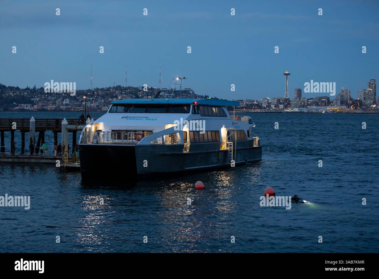 Passagiere steigen am Seacrest Ferry Dock an Bord des West Seattle Water Taxi auf dem Weg in die Innenstadt von Seattle. Im Vordergrund tauchen zwei Nachttaucher auf Stockfoto