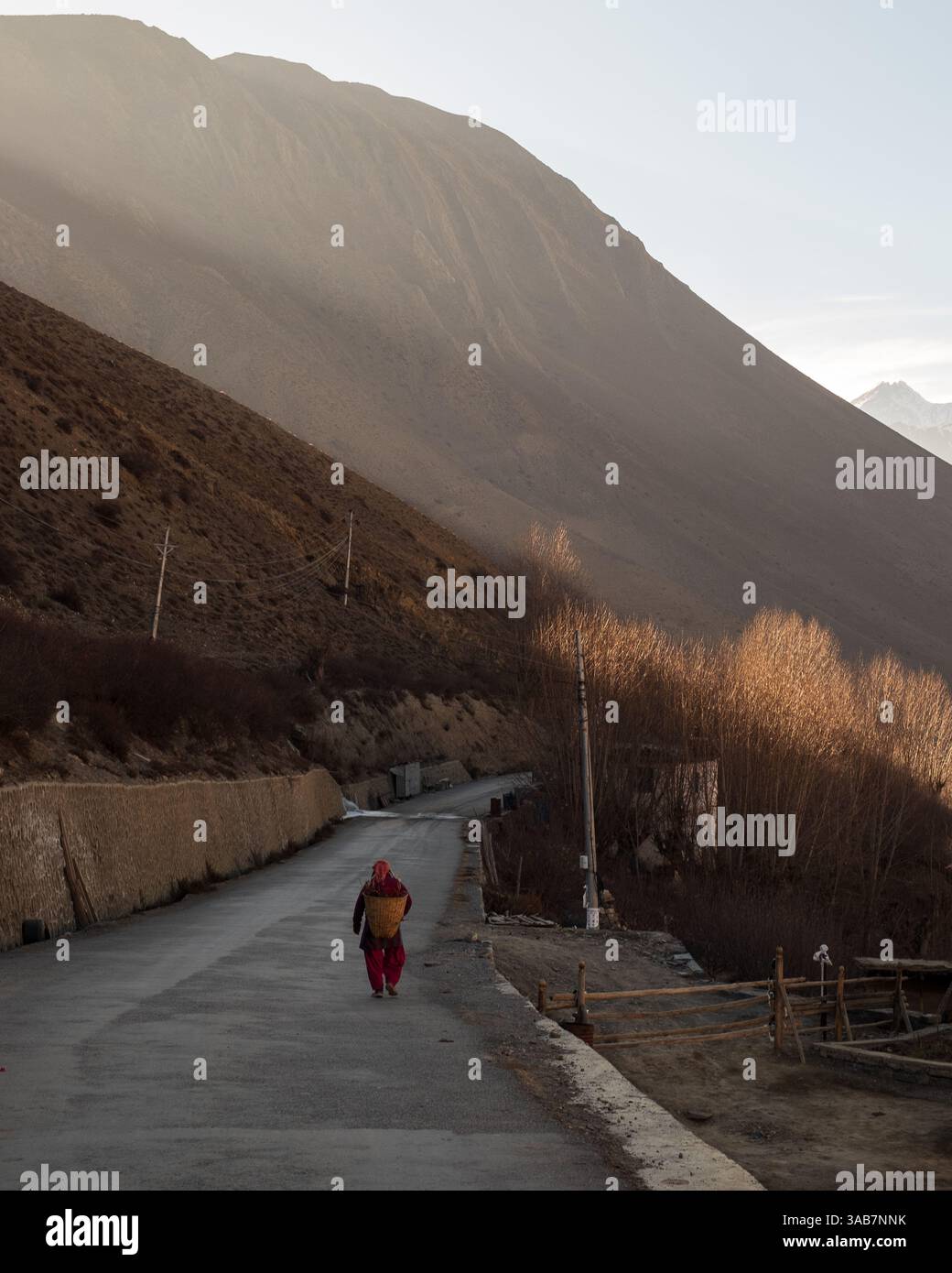 Eine nepalesische Landwirtin spaziert entlang einer asphaltierten Straße in Muktinath, Nepal, und trägt einen gewebten Korb, während die Sonne über der atemberaubenden Berglandschaft untergeht. Stockfoto