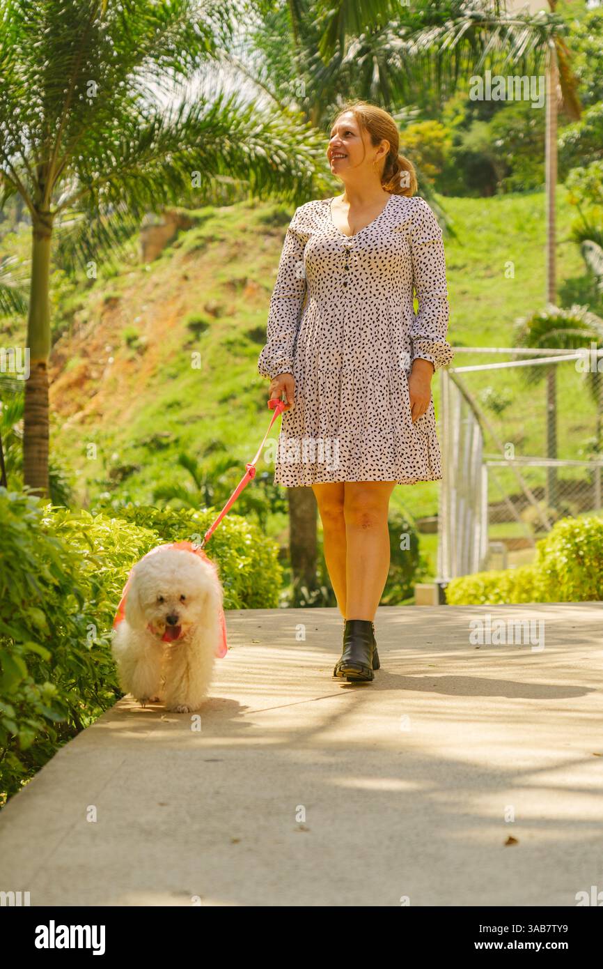 Glückliche reife Frau in einem Sommerkleid, die ihren kleinen weißen Hund an der Leine in einem wunderschönen tropischen Park führt und einen sonnigen Tag im Freien genießt Stockfoto