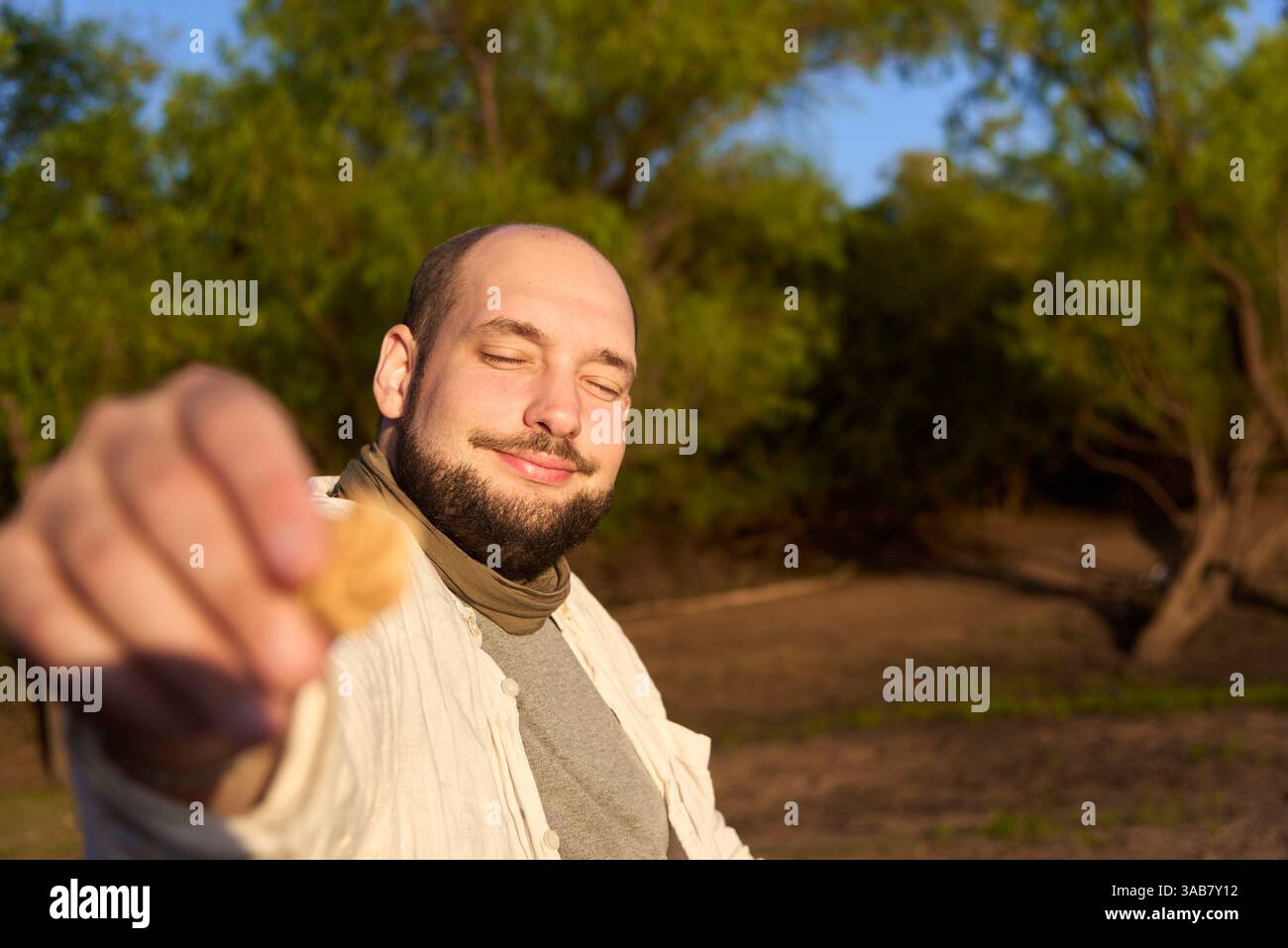 Der Mann lächelt und bietet der Kamera draußen einen Keks an. Komposition mit selektivem Fokus auf das Gesicht. Stockfoto