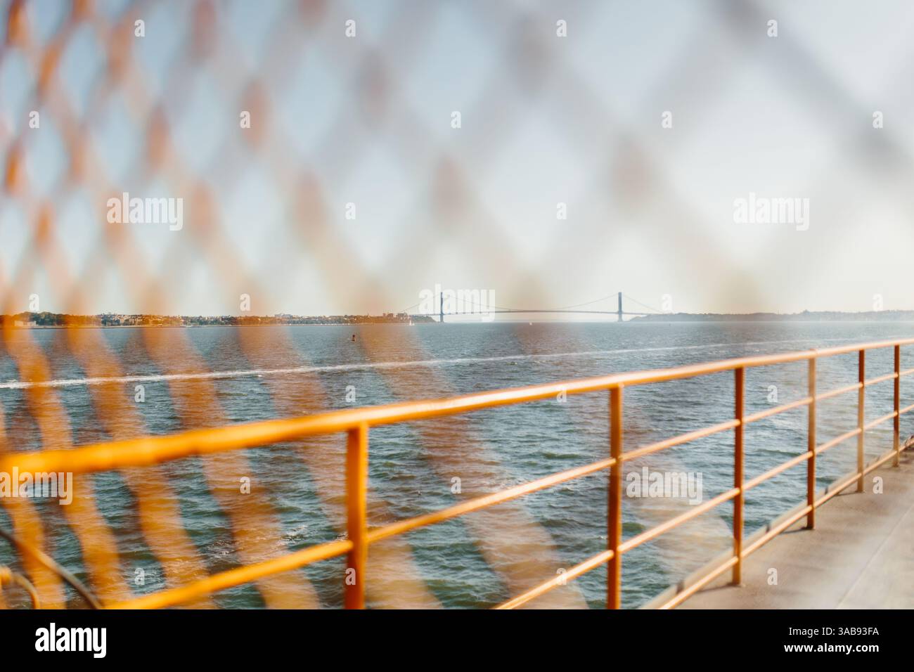 Blick von der Sonne auf eine ferne Brücke über eine ruhige Wasserstraße, gesehen durch das verschwommene Muster eines Maschendrahtzauns, mit einem orangen Geländer, das die hervorhebt Stockfoto