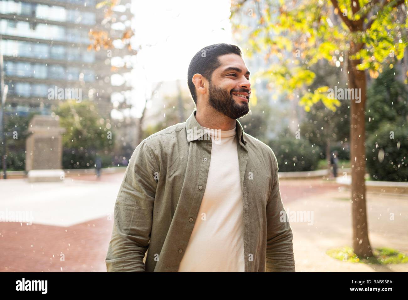 Ein Mann mit einem Bart lächelt, während er einen leichten Regen in einem Park der Stadt genießt. Er trägt eine leichte Jacke, umgeben von Bäumen und modernen Gebäuden, Captur Stockfoto