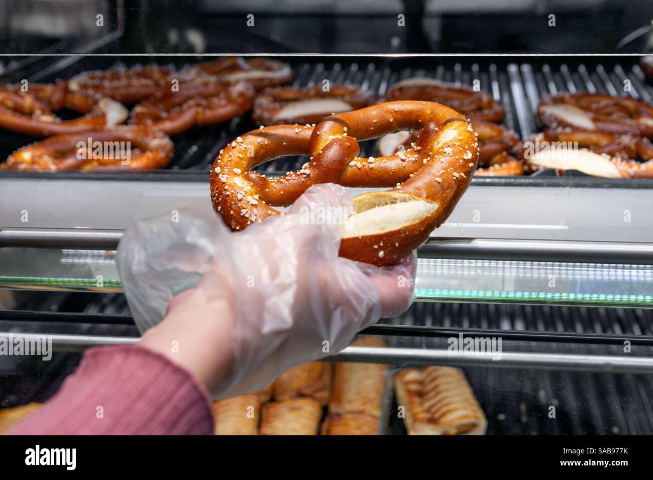Eine Nahaufnahme einer frisch gebackenen, gesalzenen Brezel, die mit Handschuhen aus einem Backwarenregal geholt wird. Die goldbraune Kruste ist mit Salz bestreut, r Stockfoto