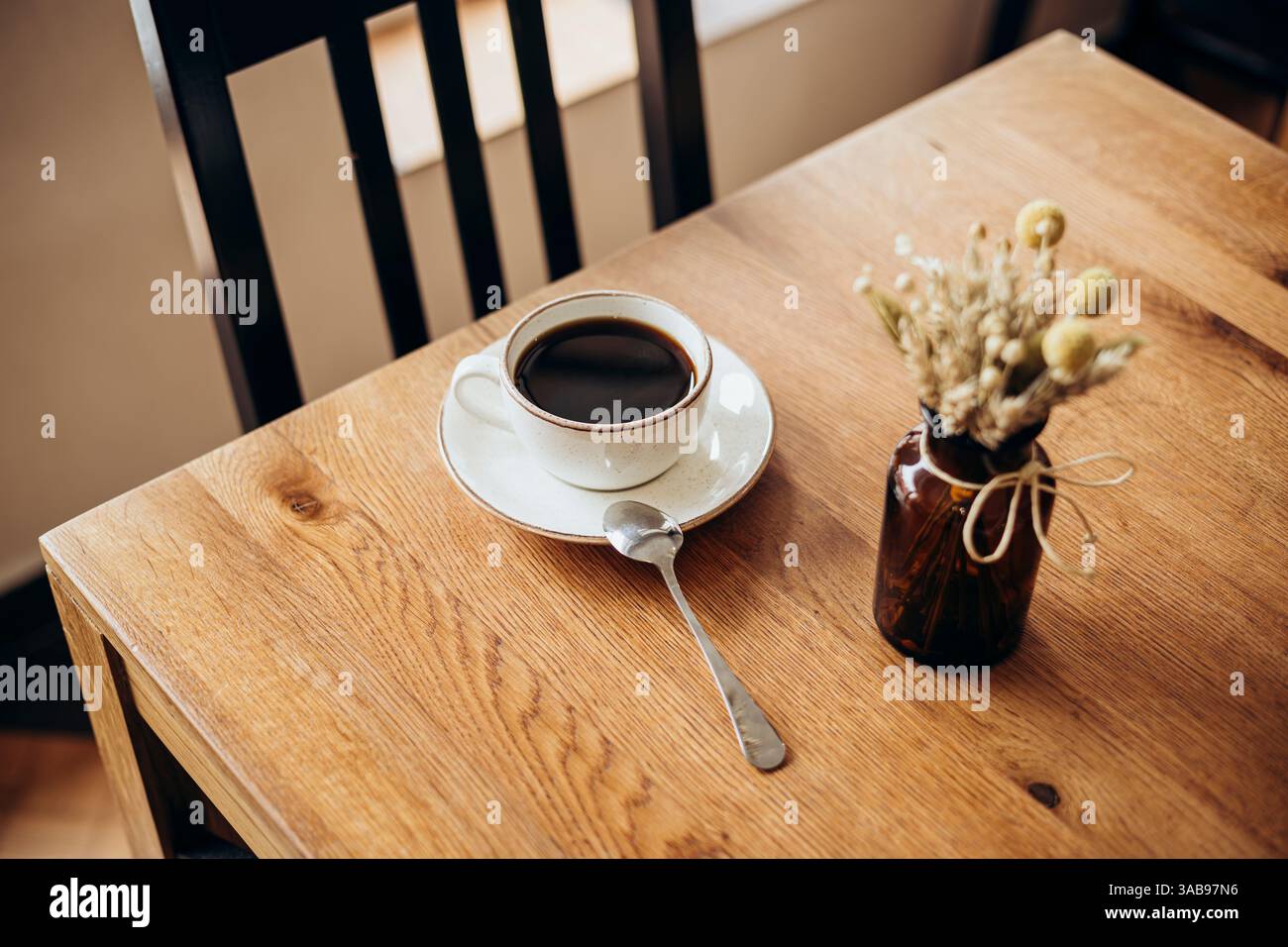 Eine gemütliche Szene mit einer Kaffeetasse auf einem Holztisch am Fenster in einem Café. Eine kleine Vase mit getrockneten Blumen verleiht einen Hauch von rustikalem Charme, perfekt für Stockfoto