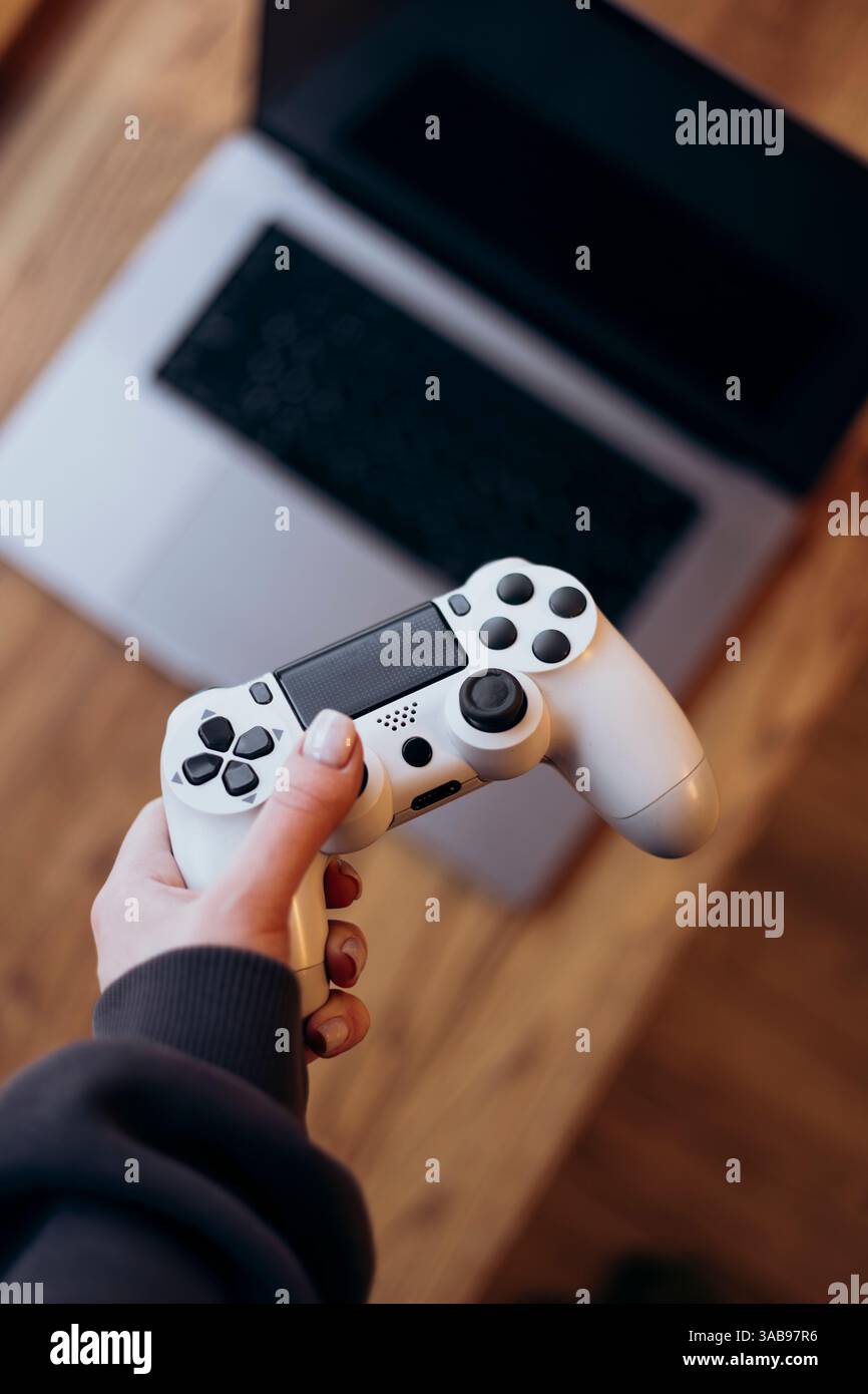 Nahaufnahme einer Person, die einen weißen Videospielcontroller mit einem Laptop im Hintergrund hält. Perfekt für die Illustration von Spielen, Technologie oder Unterhaltung Stockfoto