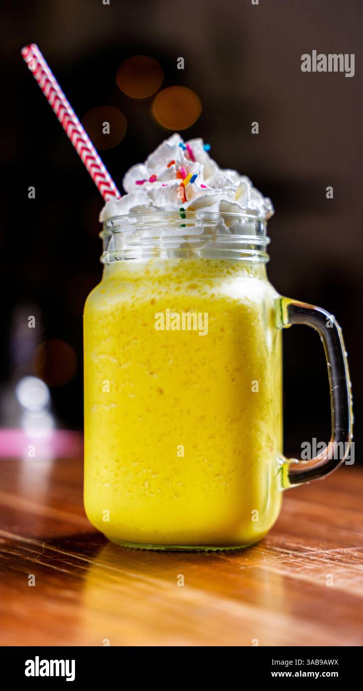 Ein lebendiger Mango-Smoothie mit cremiger Schlagsahne und bunten Streuseln, serviert in einem mauerglas mit gestreiftem Stroh auf einem hölzernen Kaffeetisch. P Stockfoto