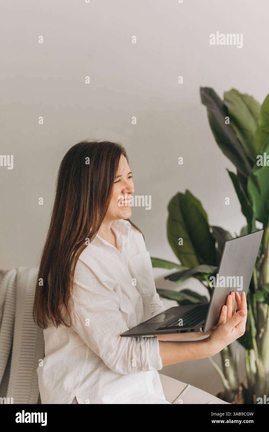 Lächelnde Ernährungsberaterin in einem weißen Hemd nimmt an einer Online-Beratung mit einem Laptop Teil. Sie sitzt an einer Pflanze und stellt eine Ruhe und Professi dar Stockfoto