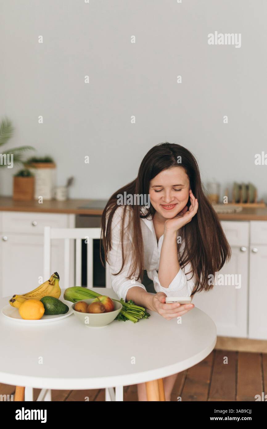 Ein Ernährungsberater ist in einer Küche, lächelt und benutzt ein Smartphone. Frisches Obst und Gemüse stehen auf dem Tisch. Die Szene vermittelt gesundes Essen und Wohlbefinden Stockfoto