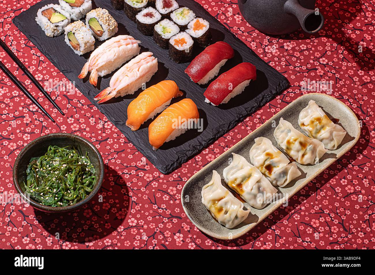 Flacher Lay eines traditionellen japanischen Essens mit Nigiri, Maki-Rollen und Gyoza, präsentiert auf einer roten Blumendecke mit dramatischen Schatten Stockfoto