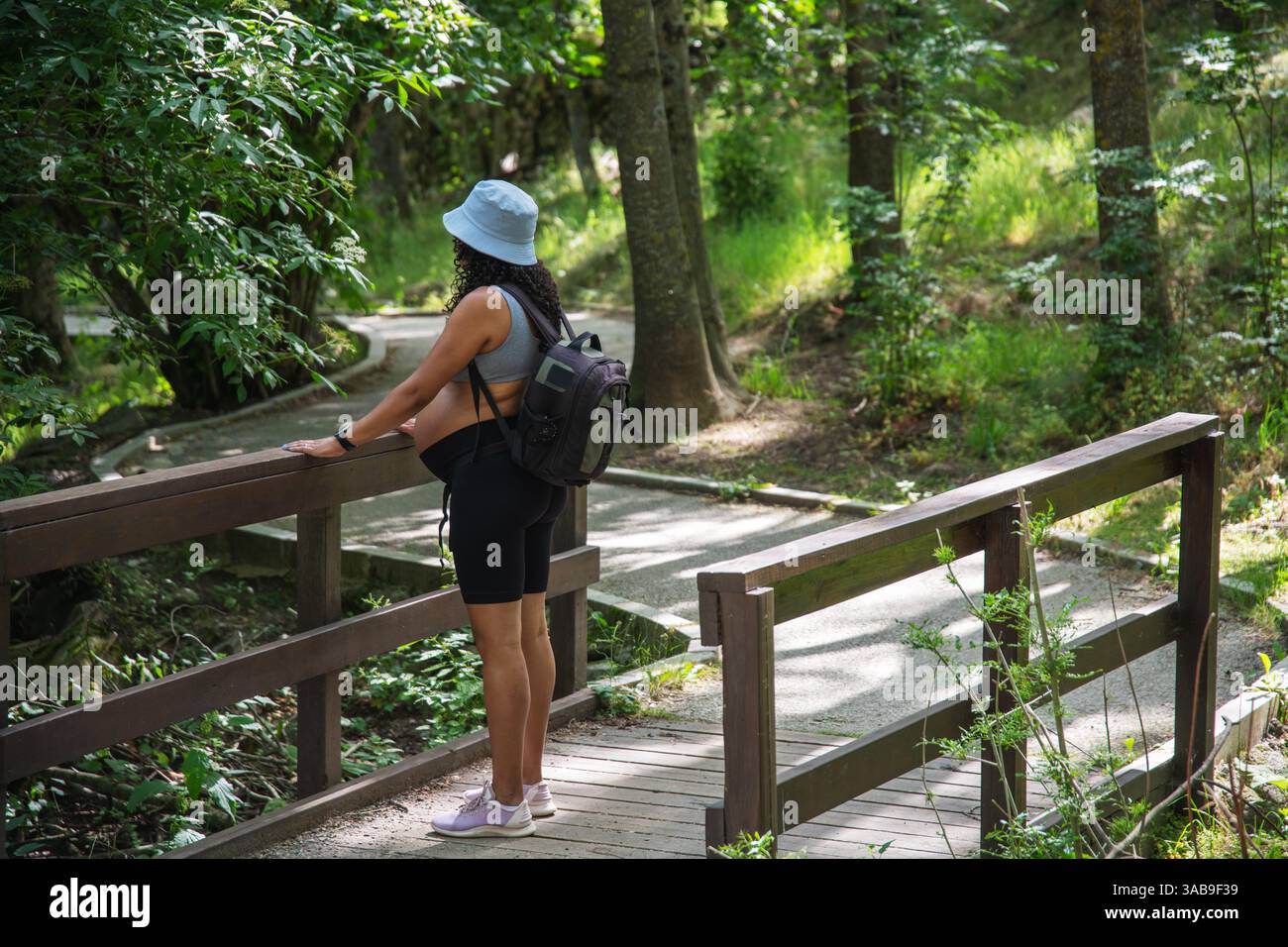 Eine schwangere Frau mit Hut und Rucksack bewundert die Waldlandschaft, während sie auf einer Holzbrücke steht. Die üppige grüne Umgebung bietet eine perfekte Umgebung Stockfoto