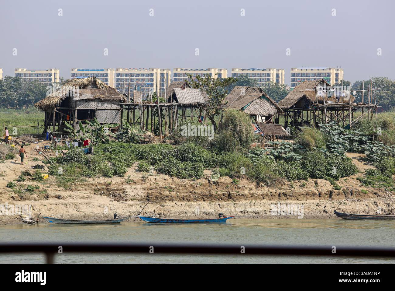 Mandalay: Irrawaddy-Ufer, traditionelle myanmarsche Pfahlhäuser, einfache umweltfreundliche Wohnungen auf Stelzen, moderner Wohnkomplex im Hintergrund Stockfoto