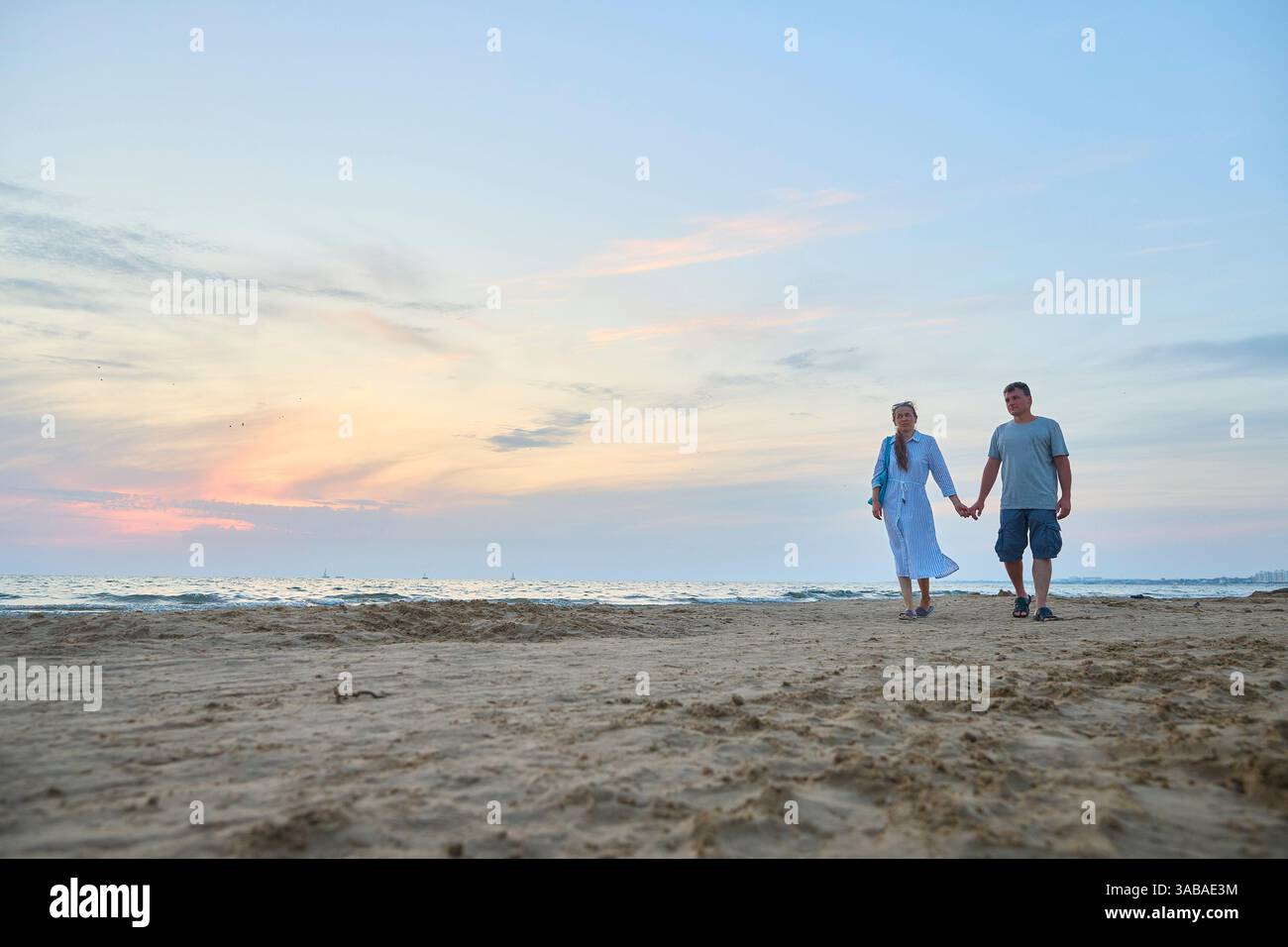 Ein paar spazieren Hand in Hand an einem Sandstrand während eines ruhigen Sonnenuntergangs entlang. Die beiden wirken fröhlich und entspannt, in der friedlichen abendlichen Atmosphäre B Stockfoto