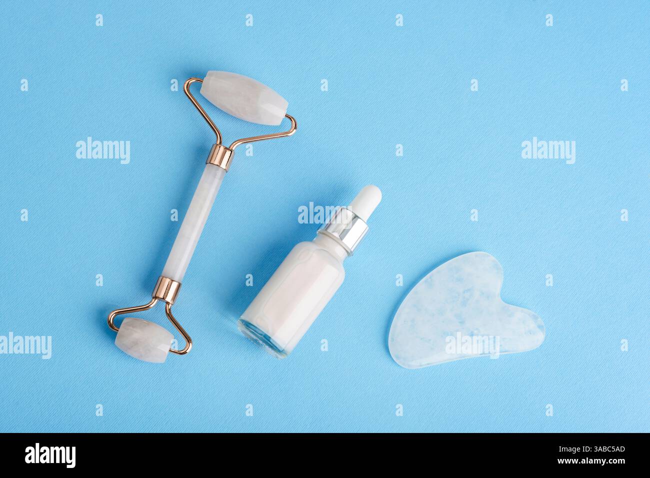 Hautpflegewerkzeuge einschließlich Gesichtsrolle, Serumtropfflasche und Gua-Sha-Stein, angeordnet auf blauem Hintergrund in minimaler, flacher Zusammensetzung für Schönheit Stockfoto