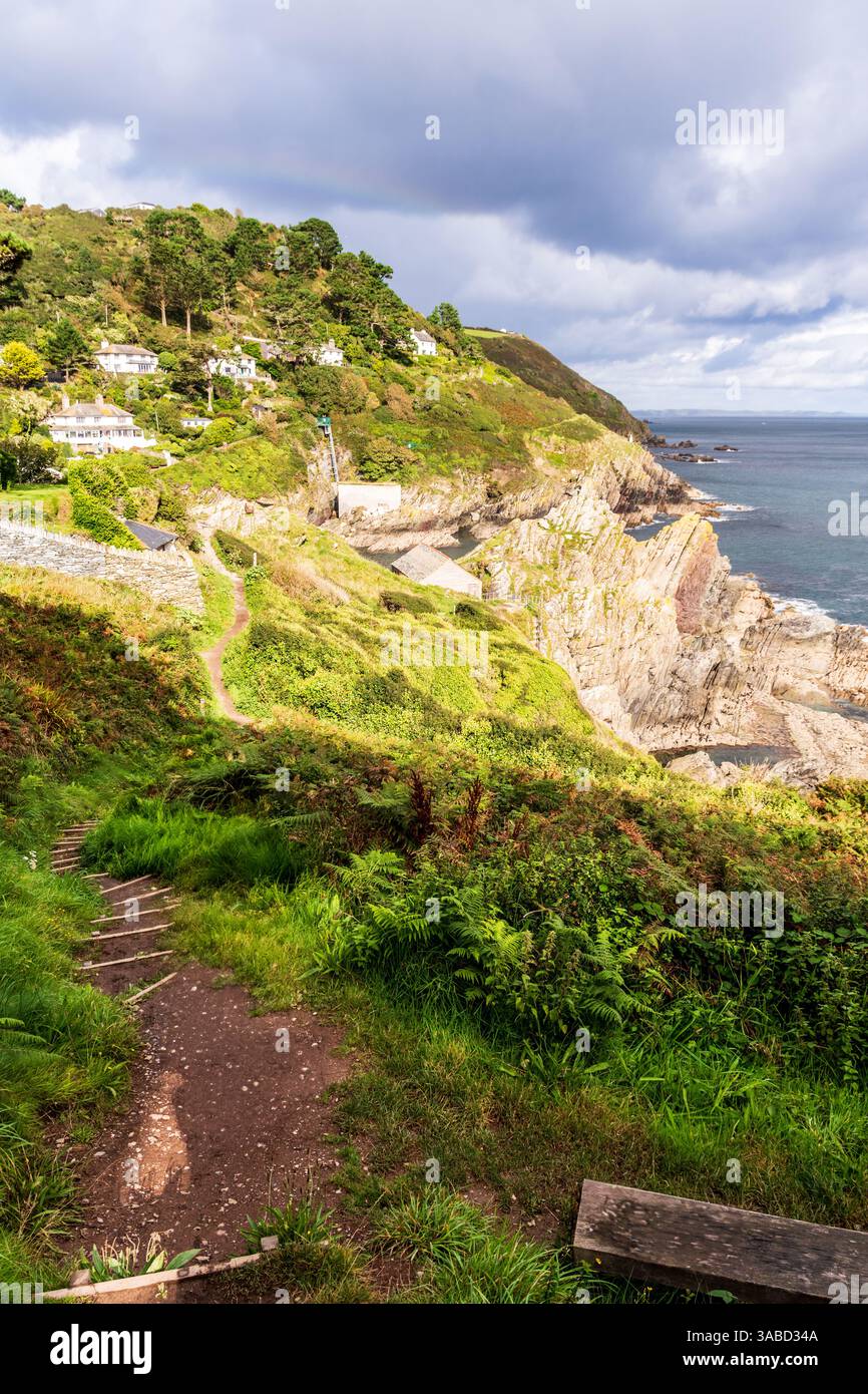 Folgen Sie dem malerischen Pfad hinunter zum bezaubernden Küstendorf Polperro, eingebettet zwischen den Klippen und dem Grün. Stockfoto