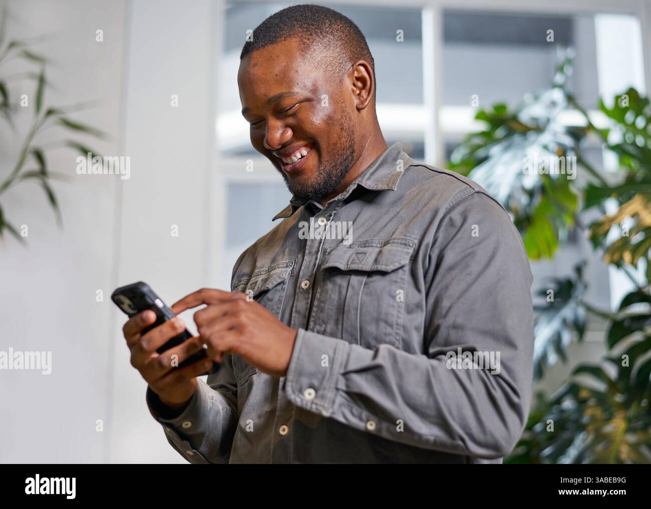 Mann lächelt, während er das Smartphone drinnen mit grünem Pflanzenhintergrund benutzt Stockfoto