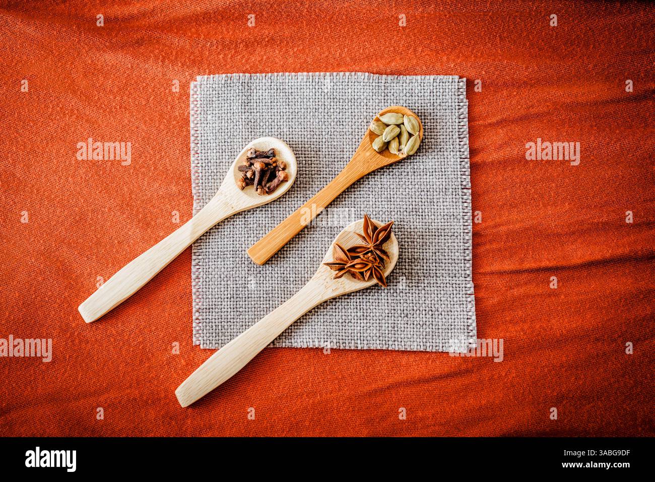 Drei Holzlöffel mit saisonalen Gewürzen auf Sackleinen-Serviette und warmen orangefarbenen Stoffen. Rustikale kulinarische Flachlage, ideal für Herbst-Food-Blogs und Bio-Co Stockfoto