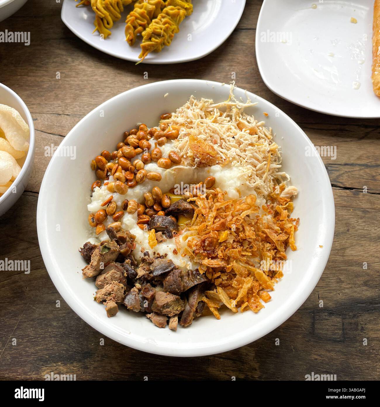 Bubur Ayam oder Hähnchenbrei, serviert mit verschiedenen Snacks auf Holztisch als Frühstückskarte - Smartphone-aufgenommenes Stockfoto