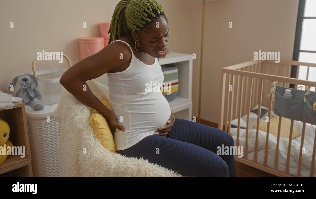 Eine Frau mit Zöpfen, die Rückenschmerzen hat, sitzt auf einem Bett in einem gemütlichen Schlafzimmer in der Nähe einer Babywiege und stellt eine heimelige und pflegende Atmosphäre dar. Stockfoto