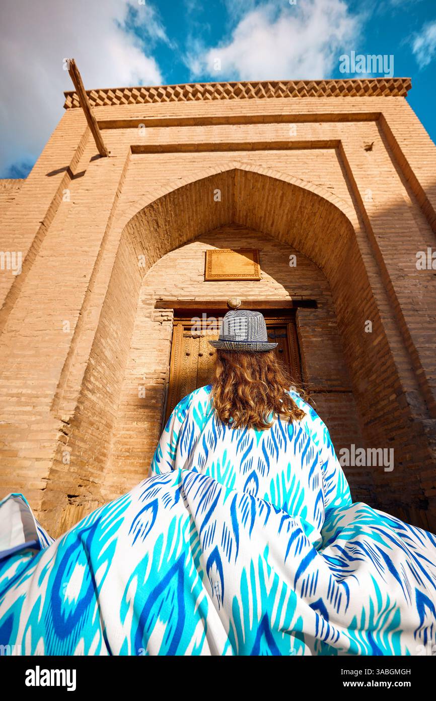 Junge Frau in ethnischer Kleidung mit blauem Ornament und Hut vor dem alten Tor an der Stadtmauer in Itchan Kala der alten Stadt Chiwa in Usbekistan Stockfoto