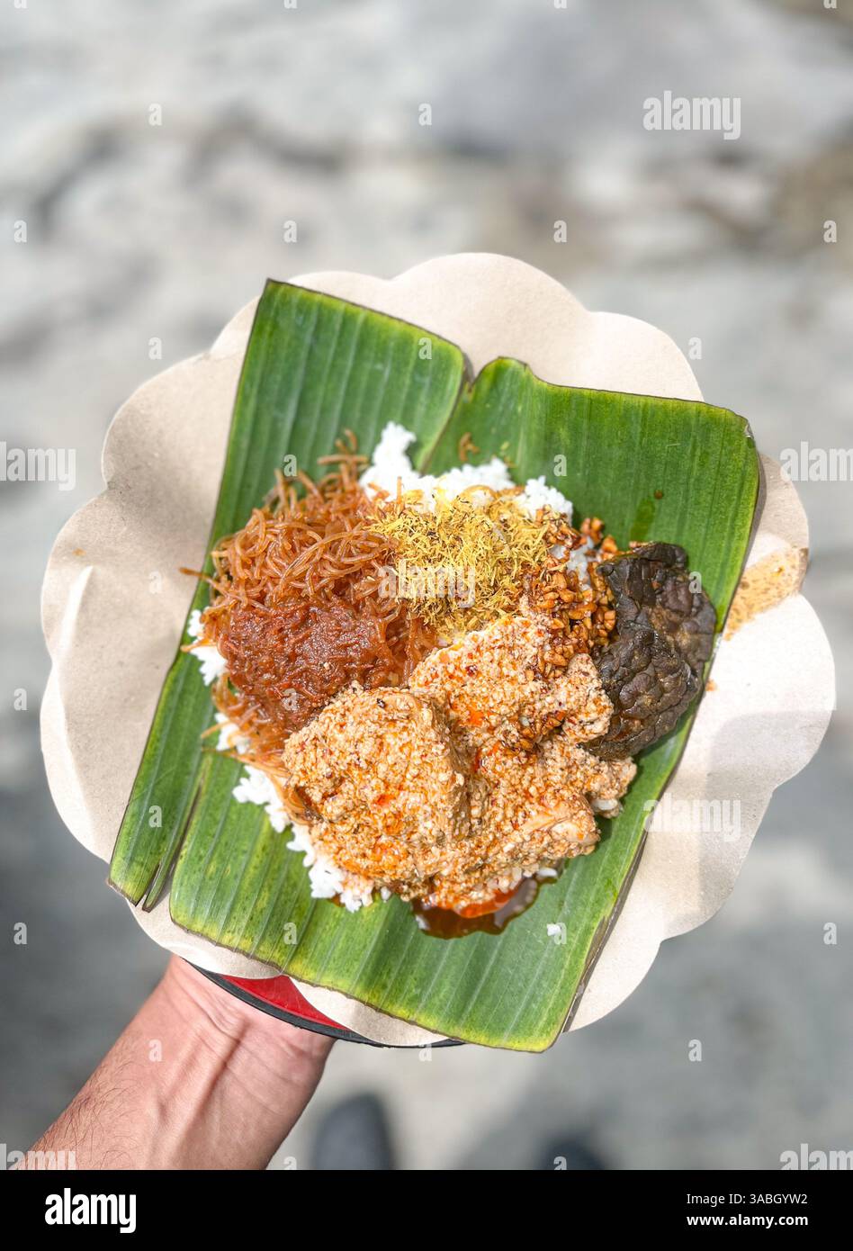 Hand hält indonesisches Essen 'nasi campur' serviert auf Teller mit Bananenblättern und unscharfem Hintergrund - Smartphone-aufgenommenes Stockfoto