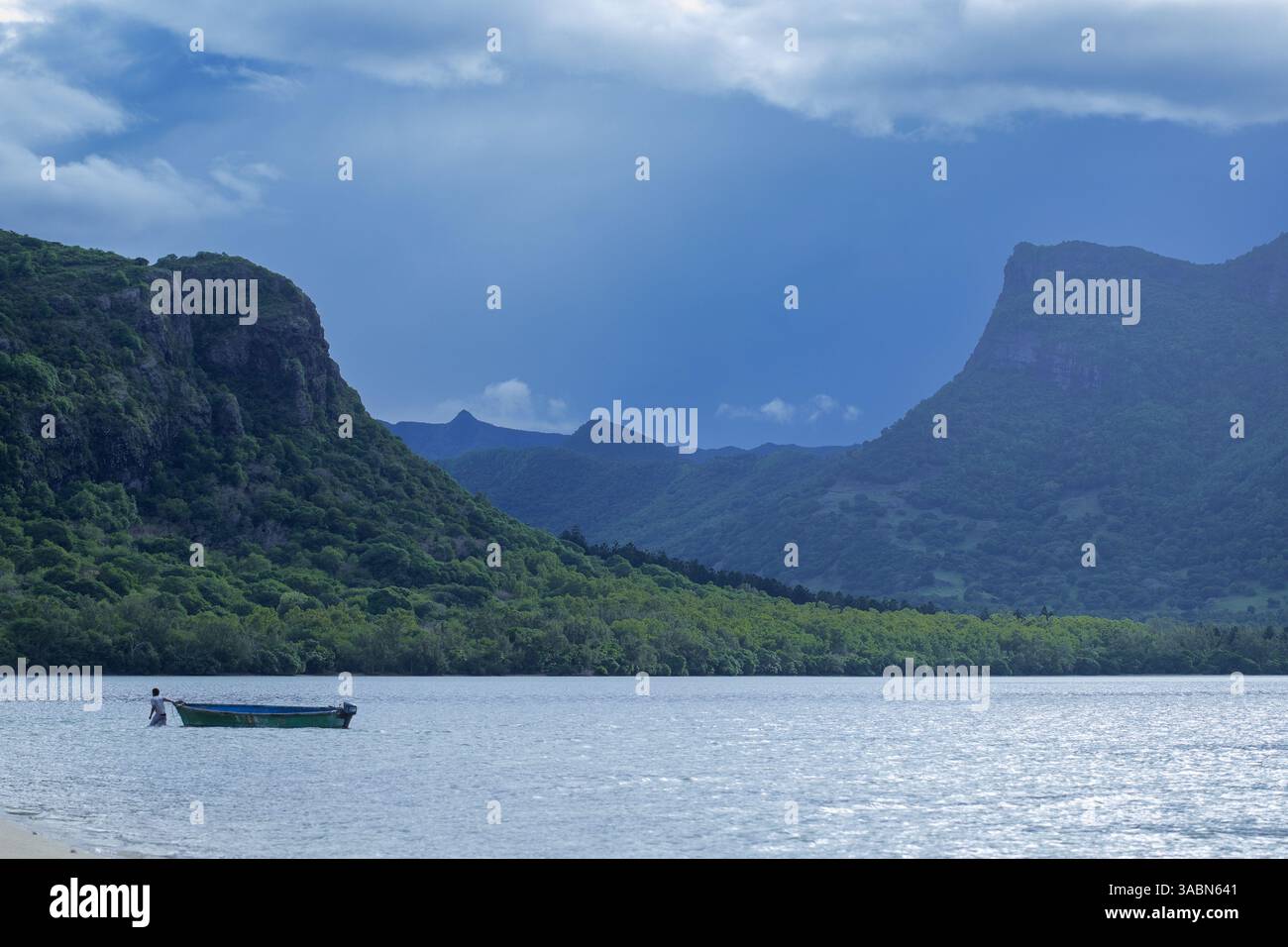 Ein Mann steht neben einem kleinen hölzernen Boot in der Nähe des Ufers, mit dem majestätischen Berg Le Morne Brabant im Hintergrund unter einem stimmungsvollen blauen Himmel Stockfoto