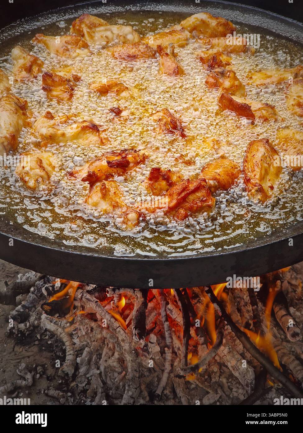 Hähnchenflügel, die draußen in einer großen Pfanne auf Feuer braten. Kochendes pflanzliches Öl lässt das Fleisch sprudeln, wenn es unter Flammen gekocht wird Stockfoto