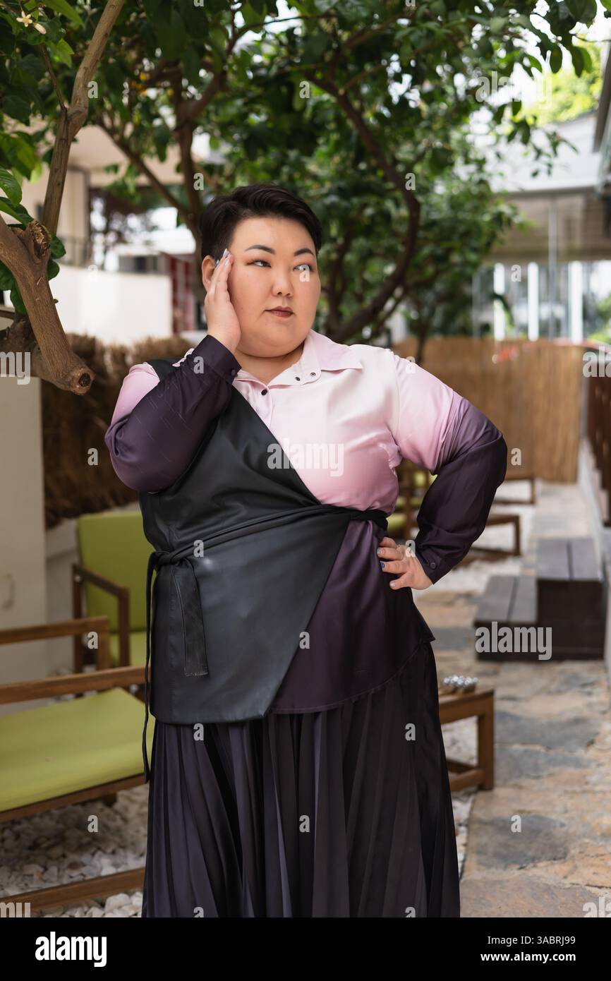 Eine junge, schöne, brünette asiatische Frau posiert mit Selbstvertrauen in einem modischen Outfit, umgeben von Grün. Körperpositiv und Lebensstil Stockfoto