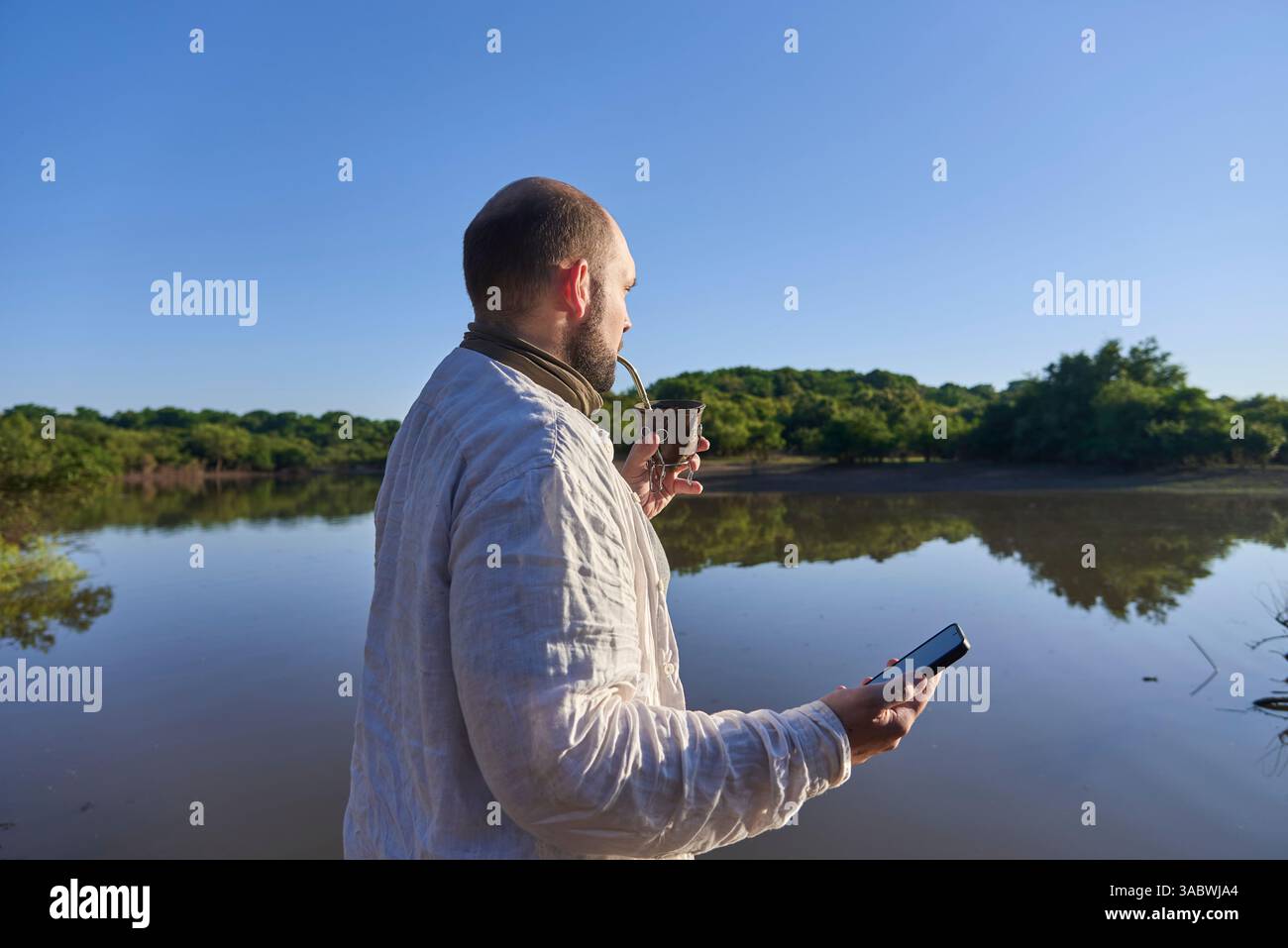 Hispanischer Mann mit Smartphone trinkt im Freien, in der Natur, blickt auf die Landschaft vor ihm, ein ruhiger Bach. El Palmar Nationalpark Stockfoto