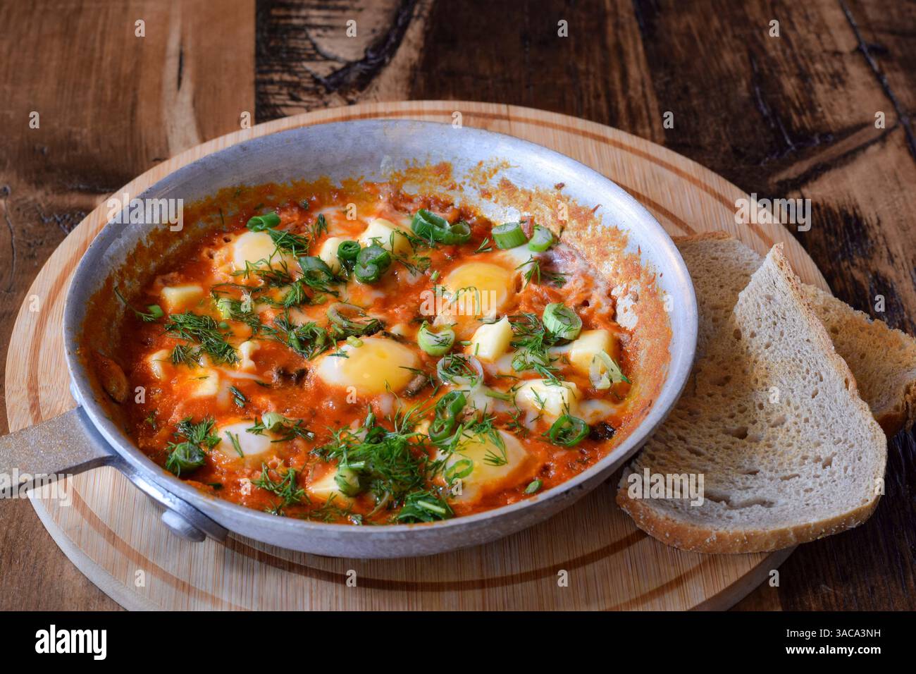 Frühstück Shakshuka garniert mit frischem Dill und grünen Zwiebeln in der Pfanne auf Holztisch mit einer Beilage Brottoast Stockfoto