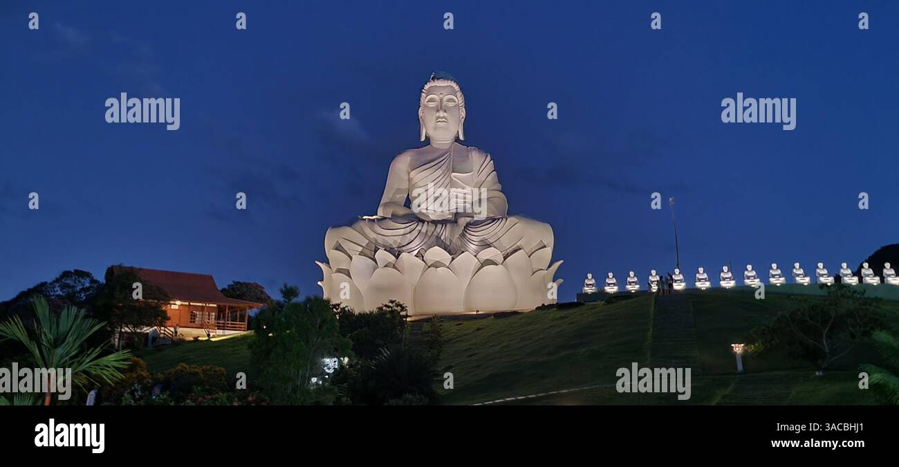 Während das letzte Licht des Tages der Dämmerung weicht, sitzt eine ruhige und majestätische weiße Buddha-Statue friedlich auf einem grünen Hügel. - Smartphone-aufgenommenes Stockfoto