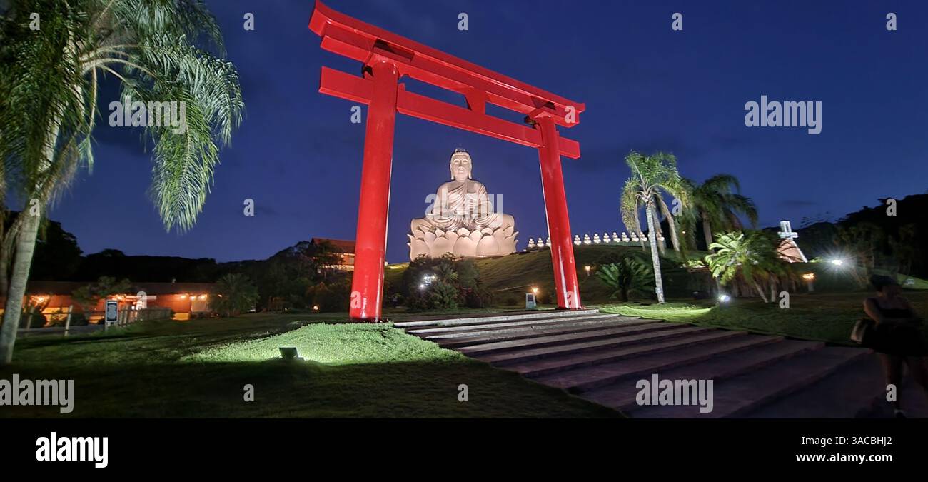 Unter dem tiefblauen Schleier der Dämmerung erhebt sich ein markantes rotes Torii-Tor, das die leuchtende Figur eines großen weißen Buddha in perfekter Harmonie umrahmt. - Smartphone-aufgenommenes Stockfoto