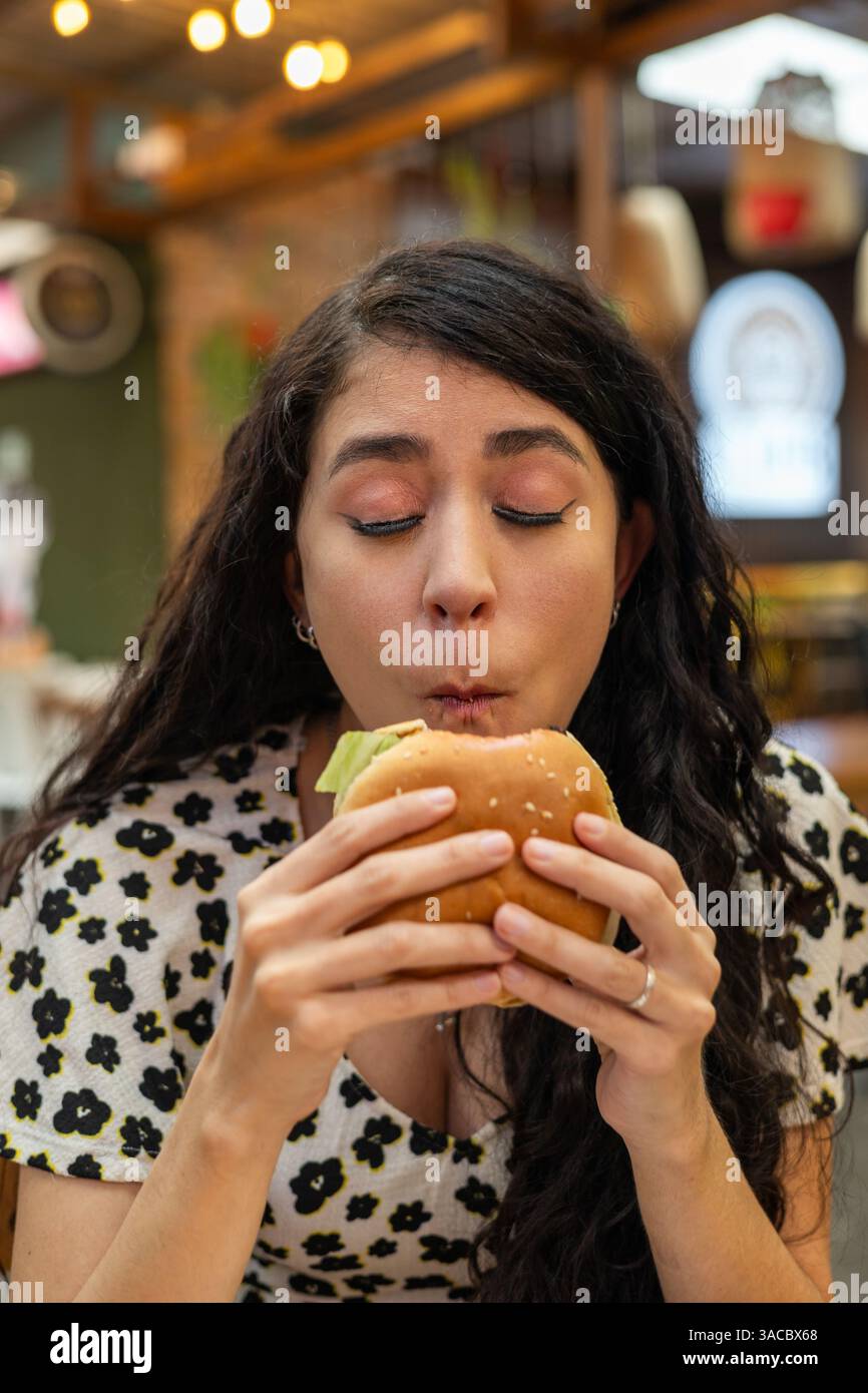 Eine junge Frau hält einen saftigen Burger mit beiden Händen und schließt ihre Augen in Erwartung eines leckeren ersten Bisses. Stockfoto
