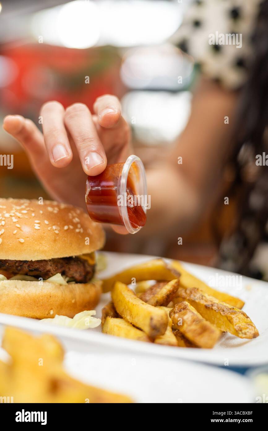 Stilvolle Frau in schwarz-weißem Kleid quetscht Ketchup auf knusprige Pommes am Restauranttisch. Authentisches Fast Food mit Burger und Getränk. Stockfoto