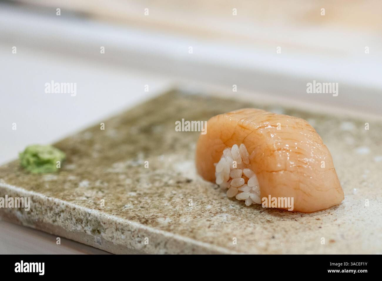 Ein einzelnes Stück Jakobsmuschel-Nigiri-Sushi liegt auf einem strukturierten Steinplatte. Das durchscheinende Fleisch glitzert im sanften Licht und zeigt seine zarte Textur. Stockfoto
