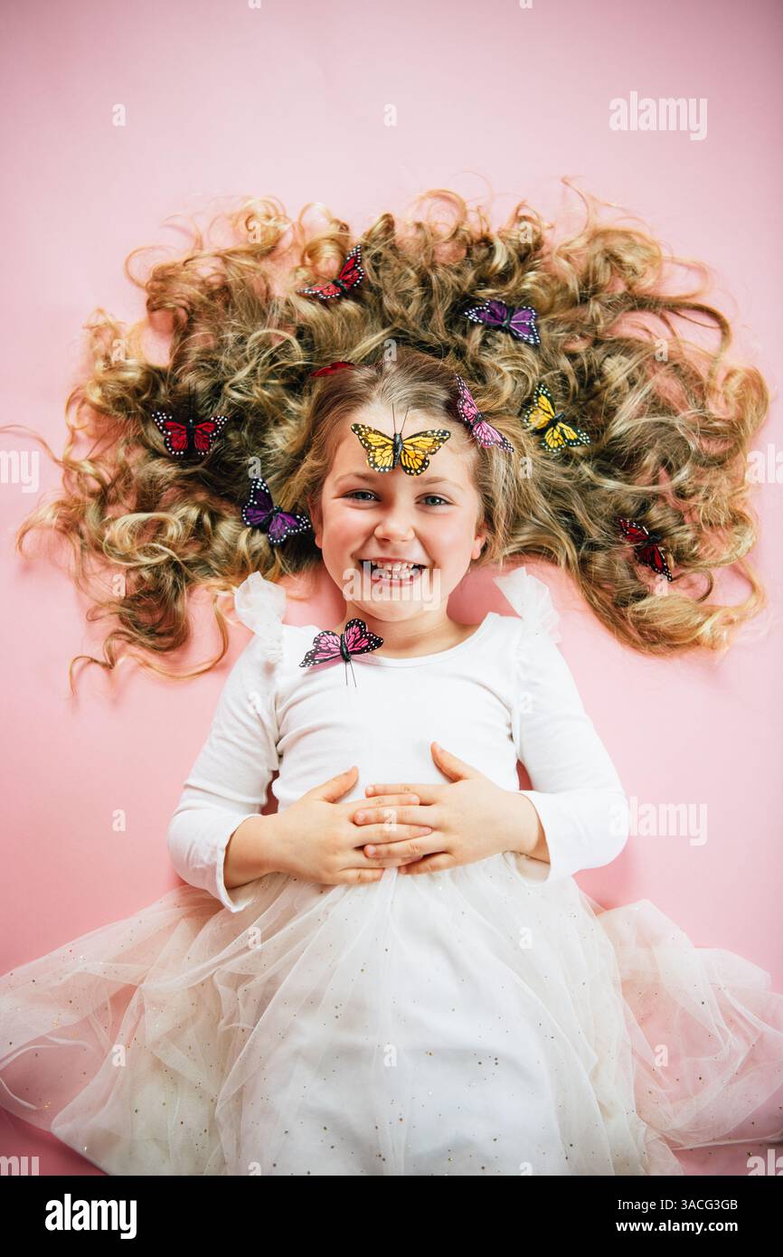 Lachendes Mädchen, das auf rosafarbenem Boden liegt, mit Schmetterlingen in lockigen Haaren Stockfoto
