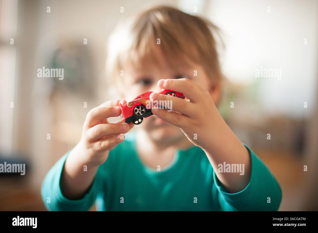 Ein Kind mit hellbraunen Haaren hält ein rotes Spielzeugauto nahe am Gesicht und lächelt in einem hellen Raum mit natürlichem Licht. Die Atmosphäre Stockfoto