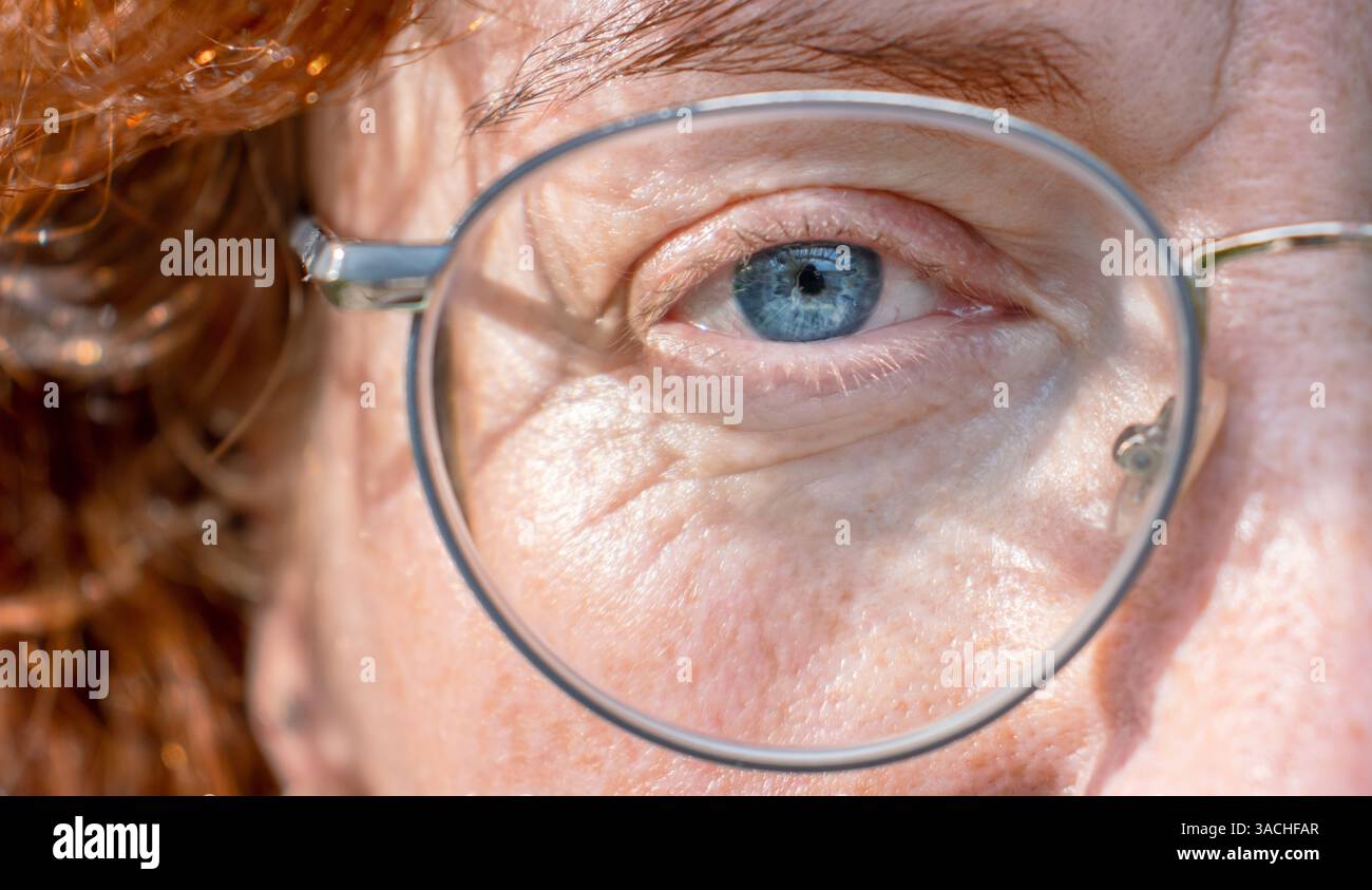 Detaillierte Nahaufnahme des rechten blaugrauen Auges einer ernsten rothaarigen Frau mittleren Alters mit einer Brille in Metallrahmen. Die Hälfte des Face Macro Shots. Fi Stockfoto