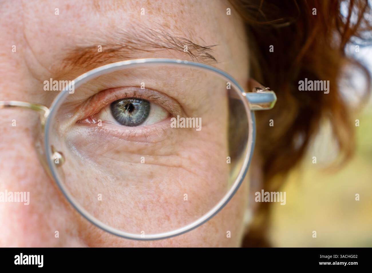 Detaillierte Nahaufnahme des linken blaugrauen Auges einer ernsten rothaarigen Frau mittleren Alters mit einer Brille in Metallrahmen. Die Hälfte des Face Macro Shots. Fi Stockfoto