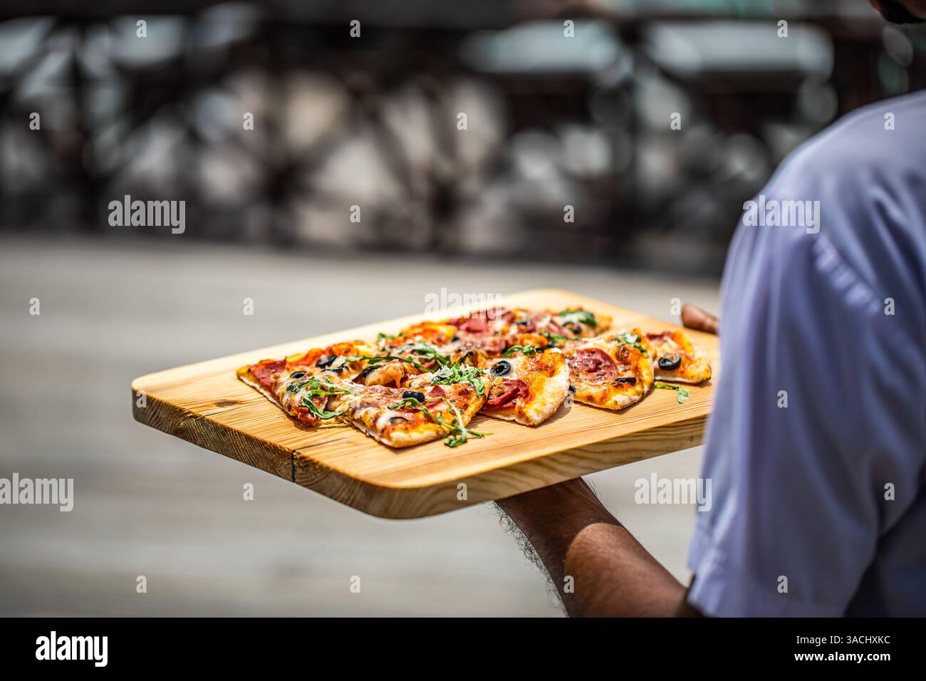 Nahaufnahme des Kellners, der Pizza zum Kunden im Restaurant im Freien brachte. Gourmet-Pizzascheiben auf Holzbrett als Teller unter Sonnenschein. Urlaubsresort Stockfoto