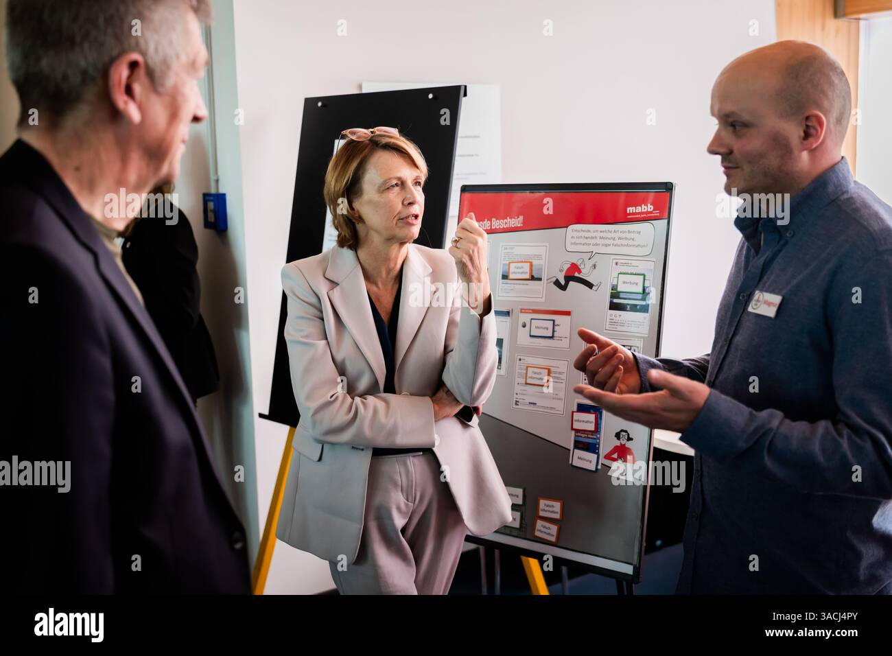 Berlin, Deutschland. April 2025. Elke Büdenbender (M), Ehefrau von Bundespräsident Steinmeier und Schirmherrin der "Nacht der Bibliotheken", spricht in der America Memorial Library am DesInfoPoint neben Volker Heller (l), Bundesvorsitzender des Deutschen Bibliotheksverbandes, mit Magnus (r) vom Medienamt Berlin-Brandenburg für Medienkompetenz und Fake News. Das Motto der vom Deutschen Bibliotheksverband (dbv) organisierten "Nacht der Bibliotheken" lautet "Wissen". Teilen. Entdecken“. Quelle: Christoph Soeder/dpa/Alamy Live News Stockfoto