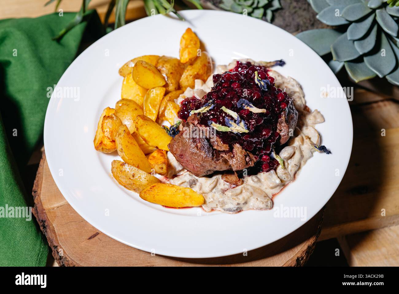 Saftiges gegrilltes Steak mit Preiselbeersauce, serviert mit gebratenen Kartoffeln und cremiger Pilzsauce auf einem weißen Teller. Stockfoto