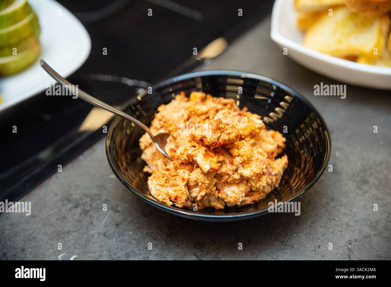 Nahaufnahme eines scharfen Käseaufstrichs mit Paprika und Kräutern, serviert in einer schwarzen Schüssel mit einem Löffel auf einer dunklen Theke. Stockfoto