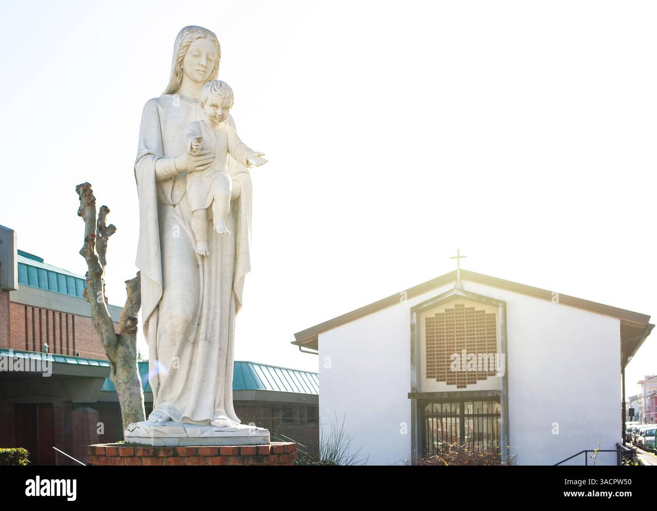 Das Sonnenlicht beleuchtet die weiße Marmorstatue der Madonna, die Kind hält, vor der modernen Kirche Stockfoto