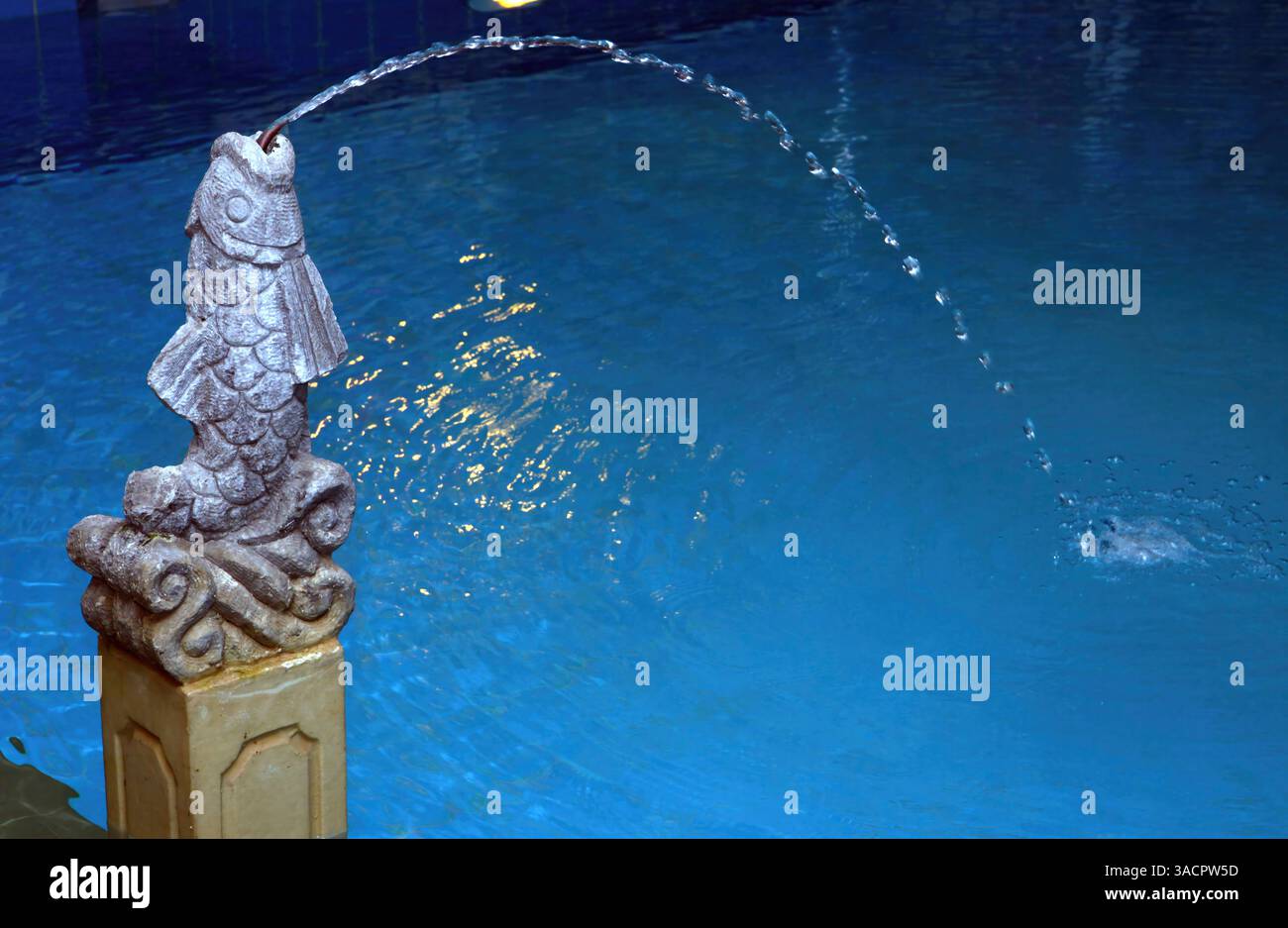 Geschwungener Wasserstrom, der anmutig aus einem Brunnen in Form eines Steinfisches sprudelt, schafft eine ruhige Atmosphäre in einem lebhaften blauen Pool Stockfoto