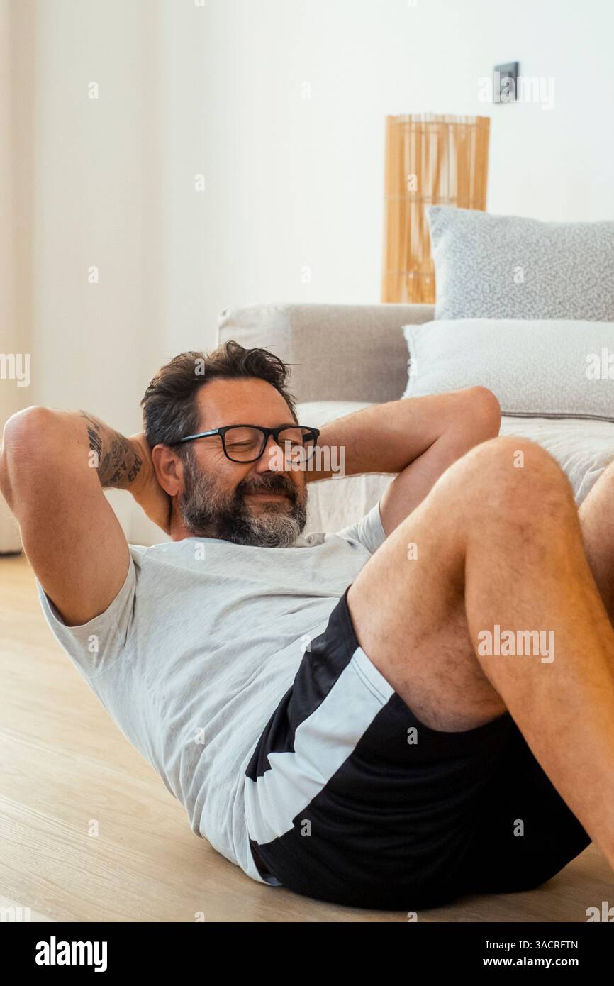 Ein erwachsener Mann von 50 Jahren, der zu Hause trainieren und Kraftübungen mit Fitnessgeräten wie Kurzhanteln und Widerstandsbändern durchführt, um sein körperliches und geistiges Wohlbefinden zu erhalten Stockfoto