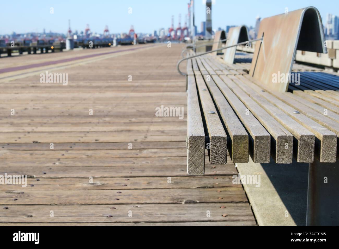 Öffentliche Parkbank mit defokussiertem Pier oder Promenade und Frachtkranen. Der Fußweg am Wasser besteht aus großen Holzdielen. Sommer Freizeit Hintergrund Textur. Stockfoto