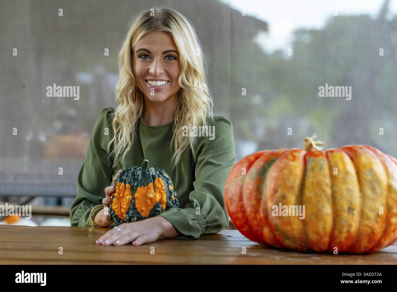 Ein atemberaubendes blondes Model posiert in einem lebhaften Kürbisfeld und trägt ein stilvolles Herbstoutfit mit einem Hauch von Halloween-Charme. Die Einstellung leuchtet warm Stockfoto