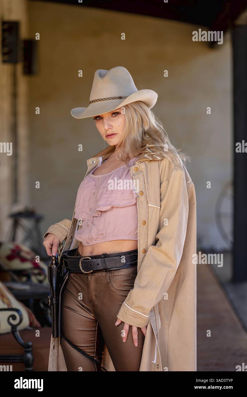 Eine selbstbewusste, schöne blonde Frau in klassischer Cowboy-Kleidung steht hoch und posiert mit einer glänzenden Pistole auf der Hüfte und einem Gewehr, das über ihrem Schuh liegt Stockfoto