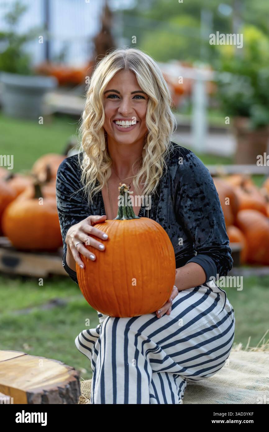 Ein atemberaubendes blondes Model posiert in einem lebhaften Kürbisfeld und trägt ein stilvolles Herbstoutfit mit einem Hauch von Halloween-Charme. Die Einstellung leuchtet warm Stockfoto
