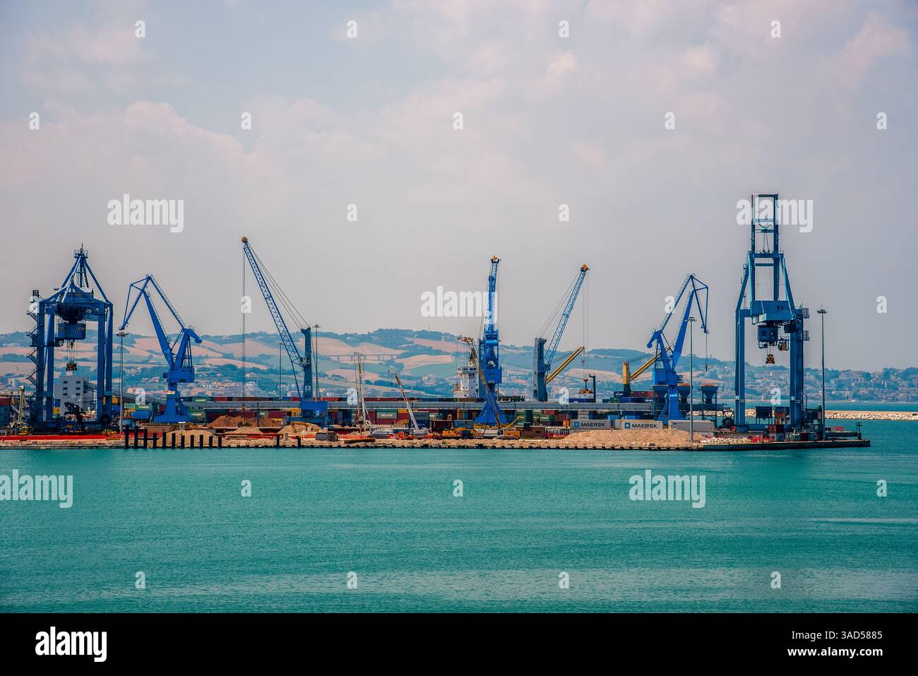 Ein Panoramablick auf einen belebten Frachthafen in Ancona, Italien, mit hoch aufragenden blauen Kränen, die sich vor sanften Hügeln der Küste und türkisfarbenem Wasser befinden Stockfoto